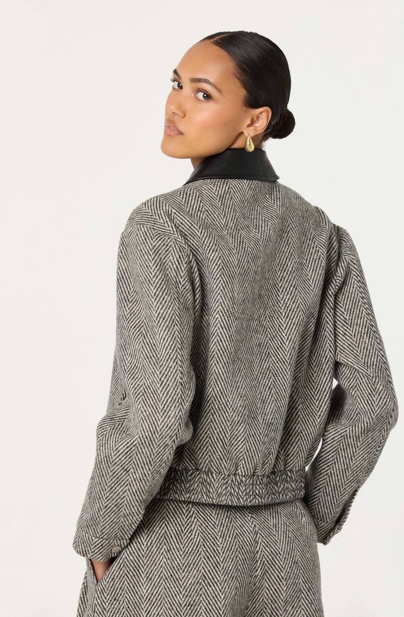 A woman is wearing a tailored jacket and matching pants made of grey and black herringbone-patterned fabric. The jacket features a black collar, and the fabric has a classic, textured appearance.