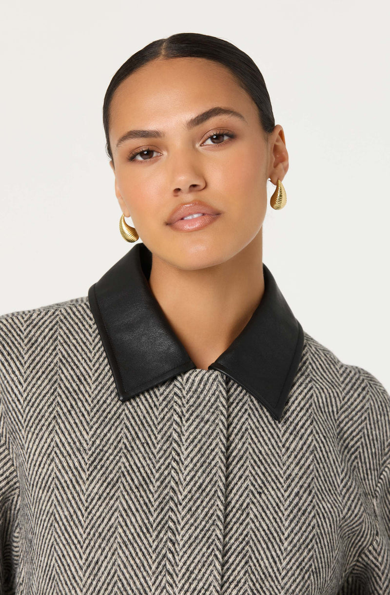 A woman is wearing a herringbone patterned coat with a black leather collar. She has gold, shell-shaped earrings and her hair is slicked back.