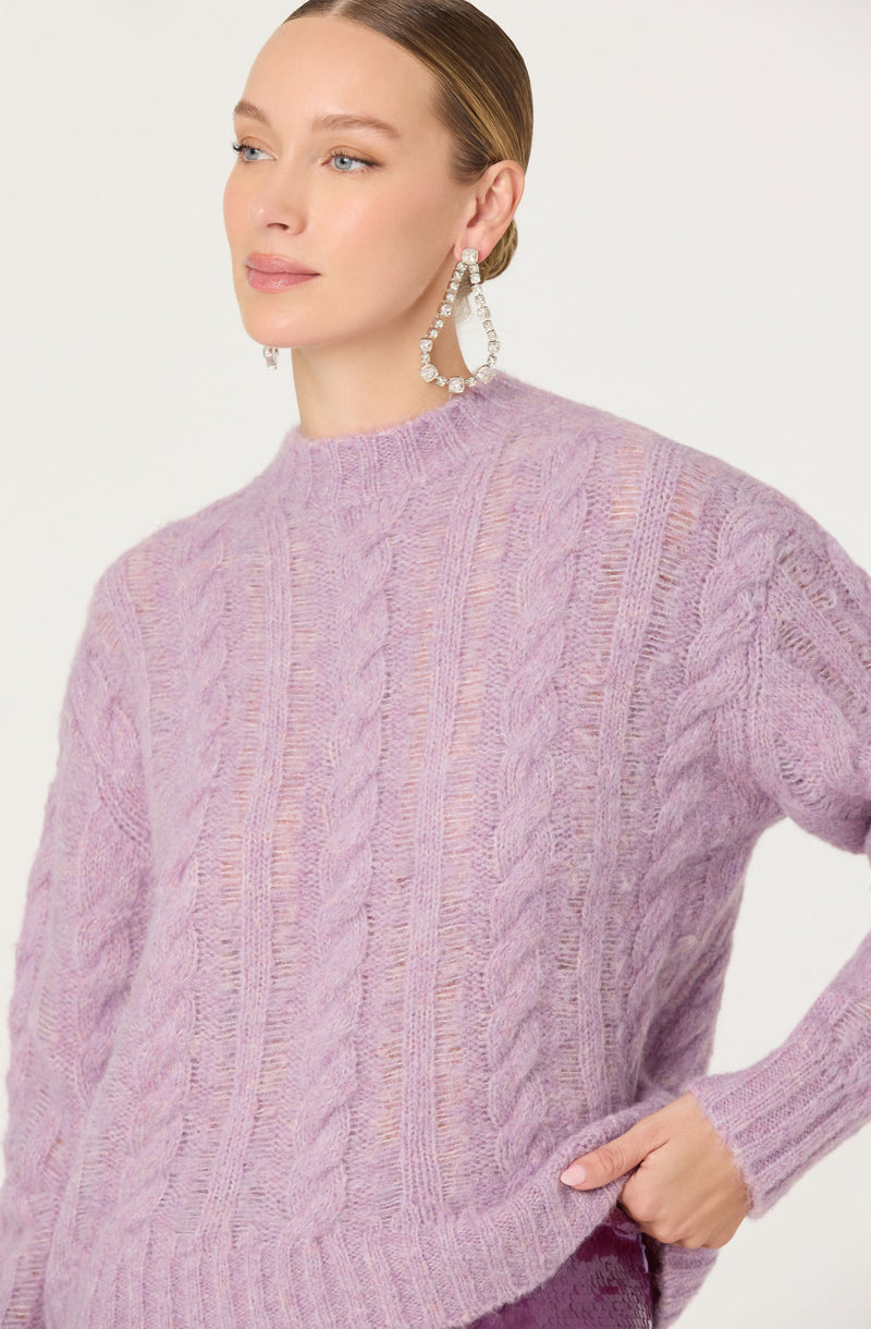 A woman is wearing a soft lavender cable-knit sweater with a high neckline. The sweater features a classic knit pattern with vertical cables running down the front and sleeves. She accessorizes with large, hoop earrings decorated with clear, sparkling stones.