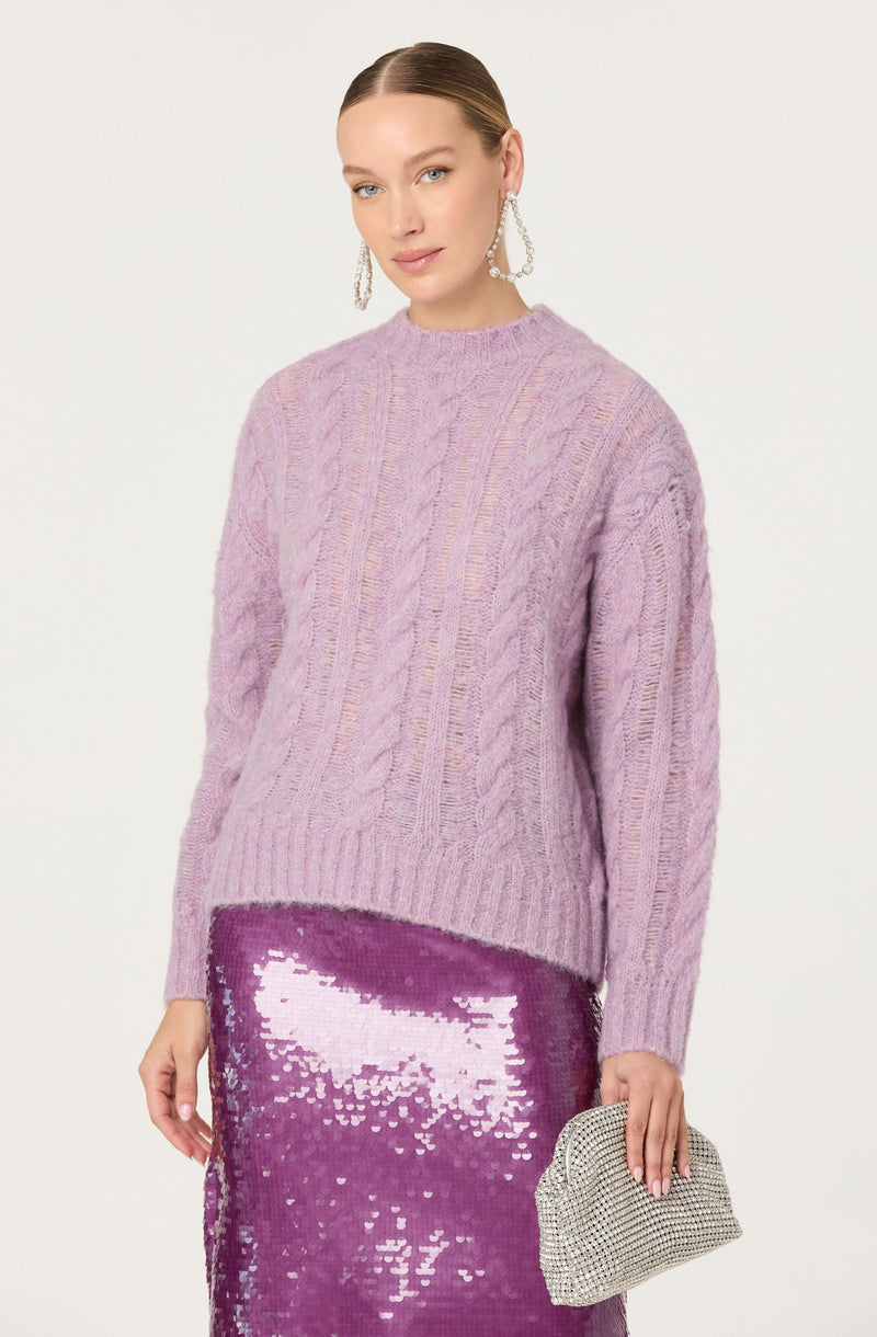 A woman is wearing a lavender-colored, chunky knit sweater with a cable knit pattern and a ribbed hem. She pairs it with a shiny, purple sequin skirt that reflects light and adds sparkle to her outfit. She accessorizes with large, hoop earrings and holds a small, silver, rhinestone-encrusted clutch bag.