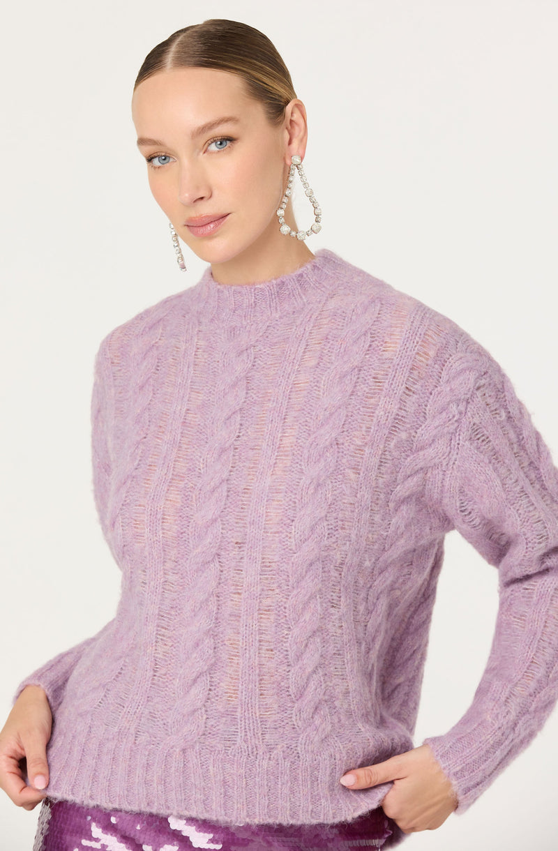 A woman is wearing a soft, oversized, light purple cable-knit sweater with a high neckline. The sweater features a textured pattern of vertical cables running down the front and sleeves. She is accessorized with large, dangling pearl and rhinestone hoop earrings.