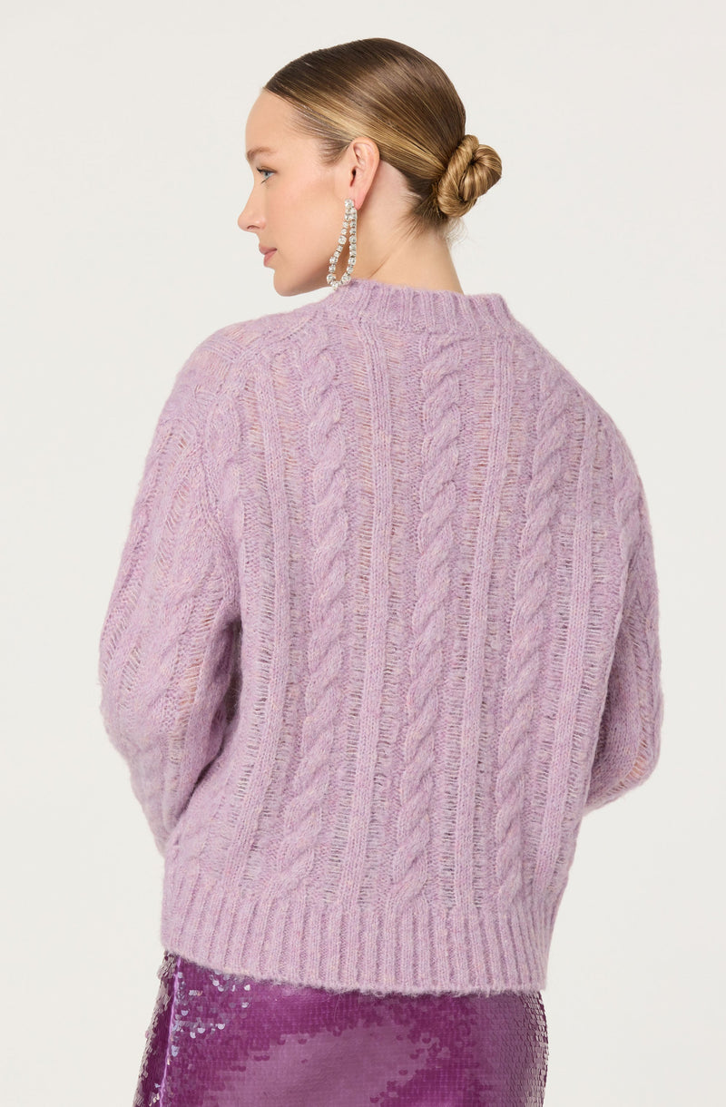A woman is wearing a light purple, chunky knit sweater with a cable-knit pattern and a high, rounded neckline. She is also wearing large, dangling, silver hoop earrings.