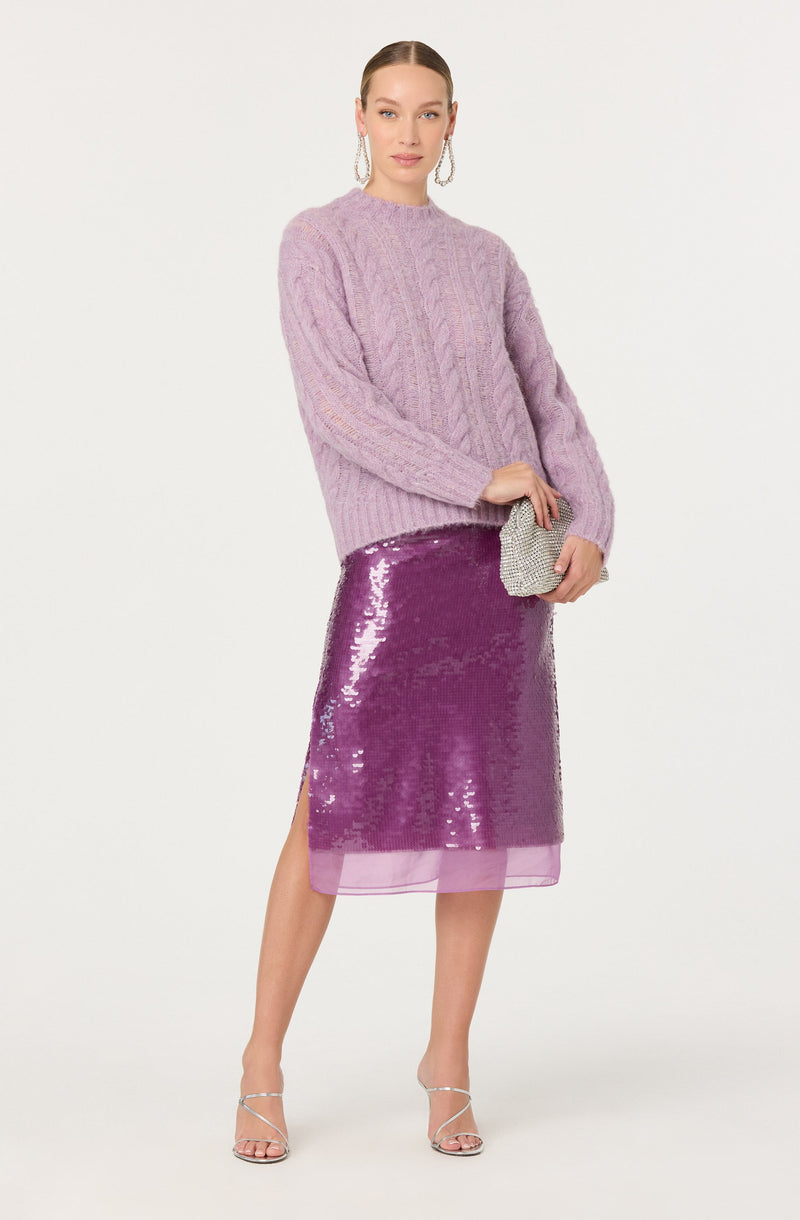 A woman is wearing a lavender cable knit sweater with a high neckline and long sleeves. She has paired it with a purple sequined skirt that has a sheer underlayer and a thigh-high slit on one side. She is holding a small, silver, rhinestone clutch and wearing strappy silver high-heeled sandals. She accessorizes with large, dangling hoop earrings.