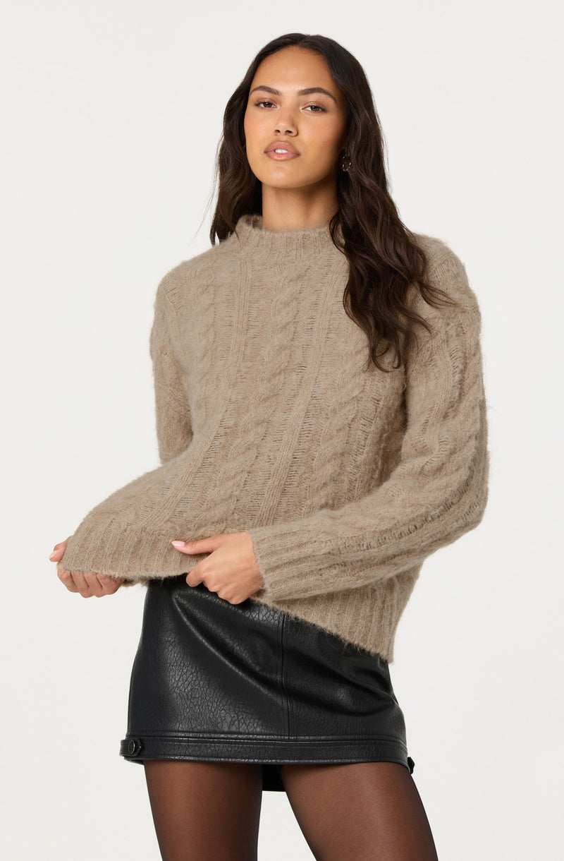 A woman is wearing a chunky, beige cable-knit sweater with a high neckline and long sleeves. She pairs it with a black faux leather mini skirt and sheer black tights.