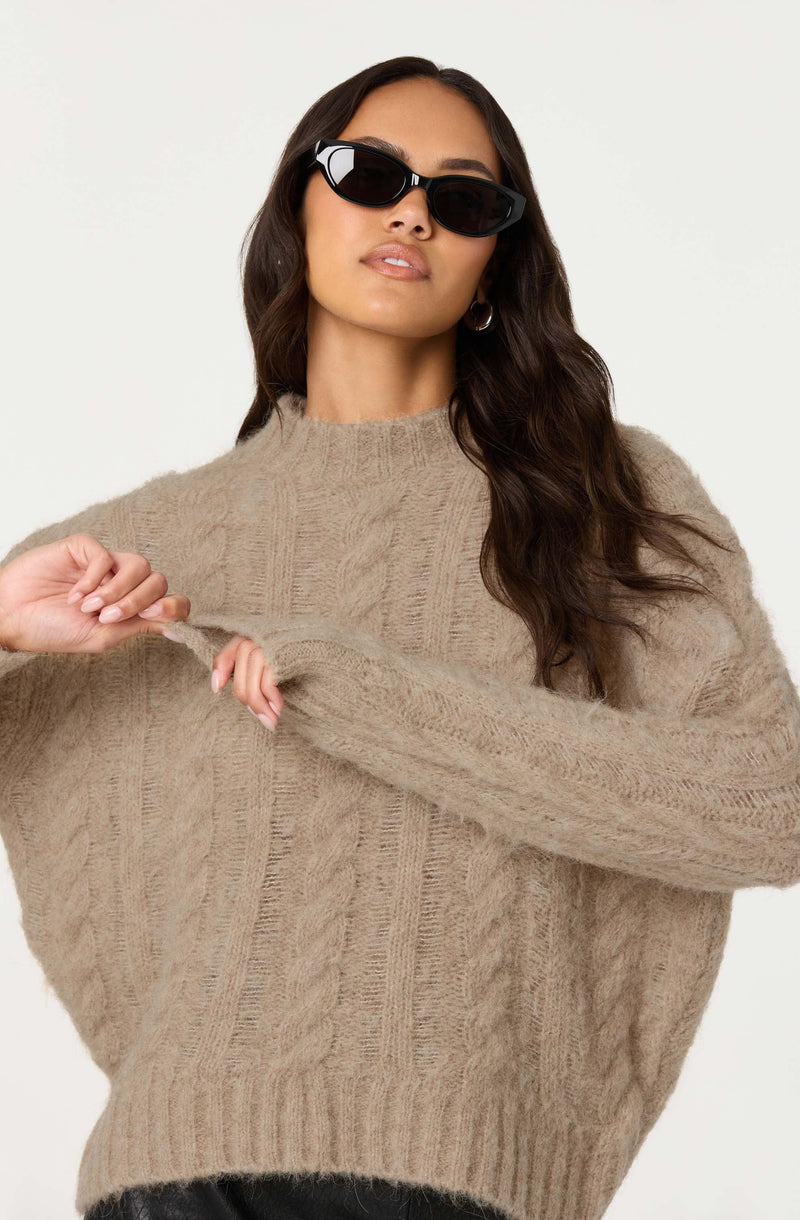 A woman is wearing oversized black sunglasses and hoop earrings. She has on a beige, chunky, cable-knit sweater with a high neckline.