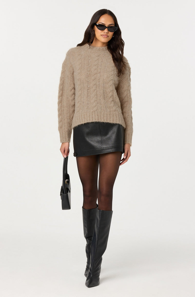 A woman is wearing a beige, chunky knit sweater with a high neckline and long sleeves. She pairs it with a black leather mini skirt, sheer black tights, and tall black leather boots that reach just below the knee. She accessorizes with black sunglasses and carries a small black handbag with a top handle.