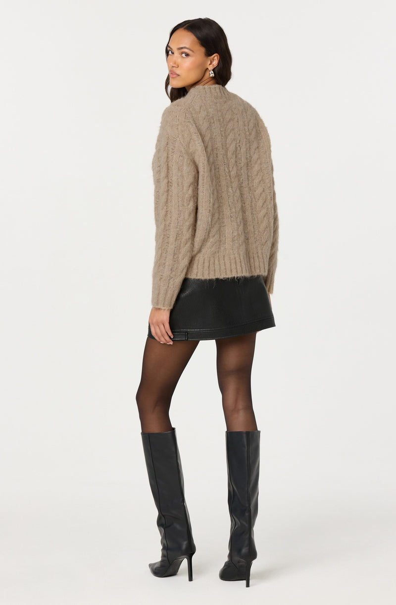 A woman is wearing a chunky, beige, cable-knit sweater with a high neckline and long sleeves. She pairs it with a black leather mini skirt, sheer black tights, and tall black leather knee-high boots with high heels.