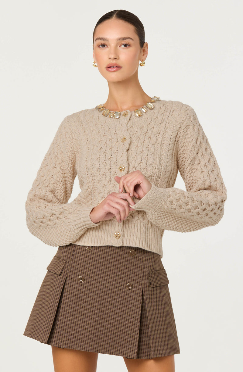 A woman is wearing a beige, cable-knit cardigan with decorative gold buttons and an embellished neckline featuring large, clear stones. She is also dressed in a brown, high-waisted plaid skirt with pleats, front pockets, and gold button details.