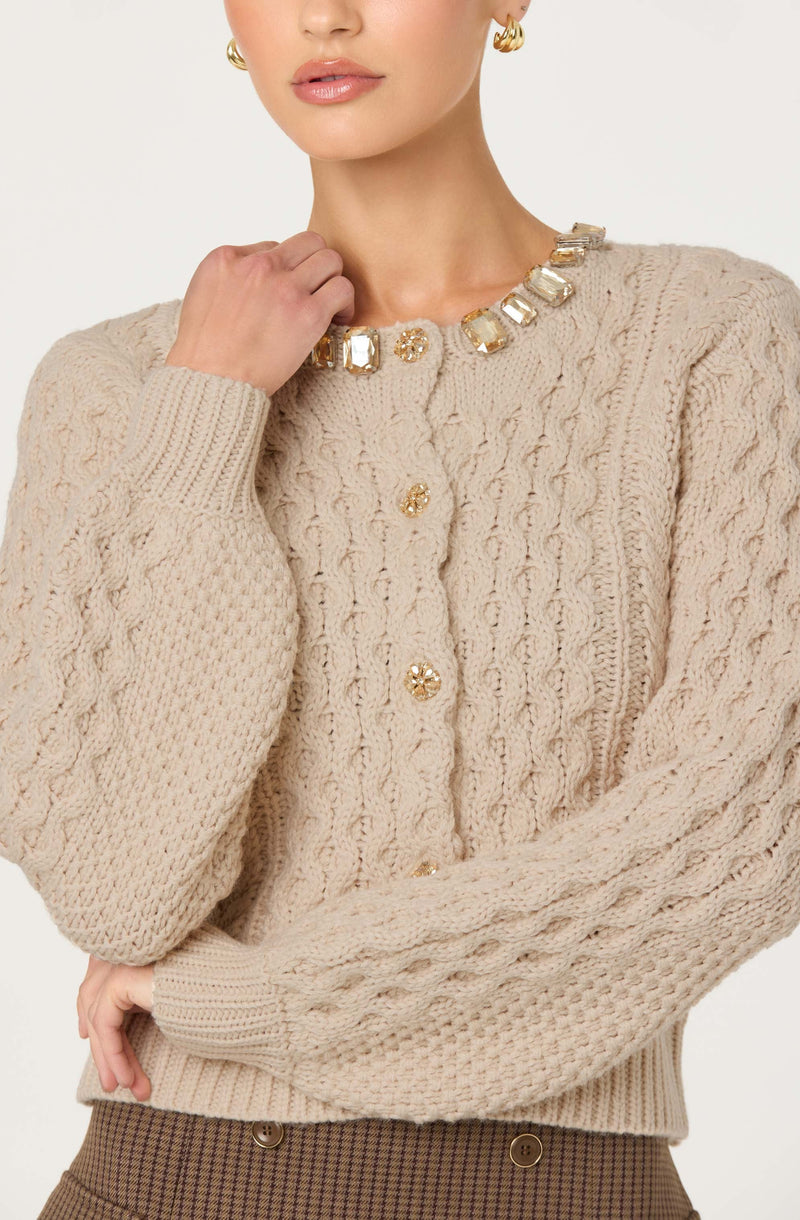 A woman is wearing a beige, cable-knit cardigan with decorative gold buttons down the front. She has accessorized with a chunky, clear statement necklace featuring large, rectangular and circular stones, and matching gold hoop earrings. The cardigan has a textured pattern with ribbed cuffs and hem.