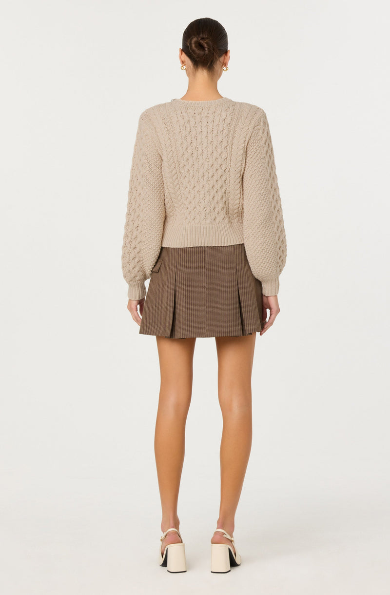 A woman is wearing a beige chunky knit sweater with textured patterns and long, puffed sleeves. She pairs it with a high-waisted, brown plaid mini skirt that features front pleats and side zipper details. She completes the outfit with white platform heels that have ankle straps. Her hair is styled in a neat bun.