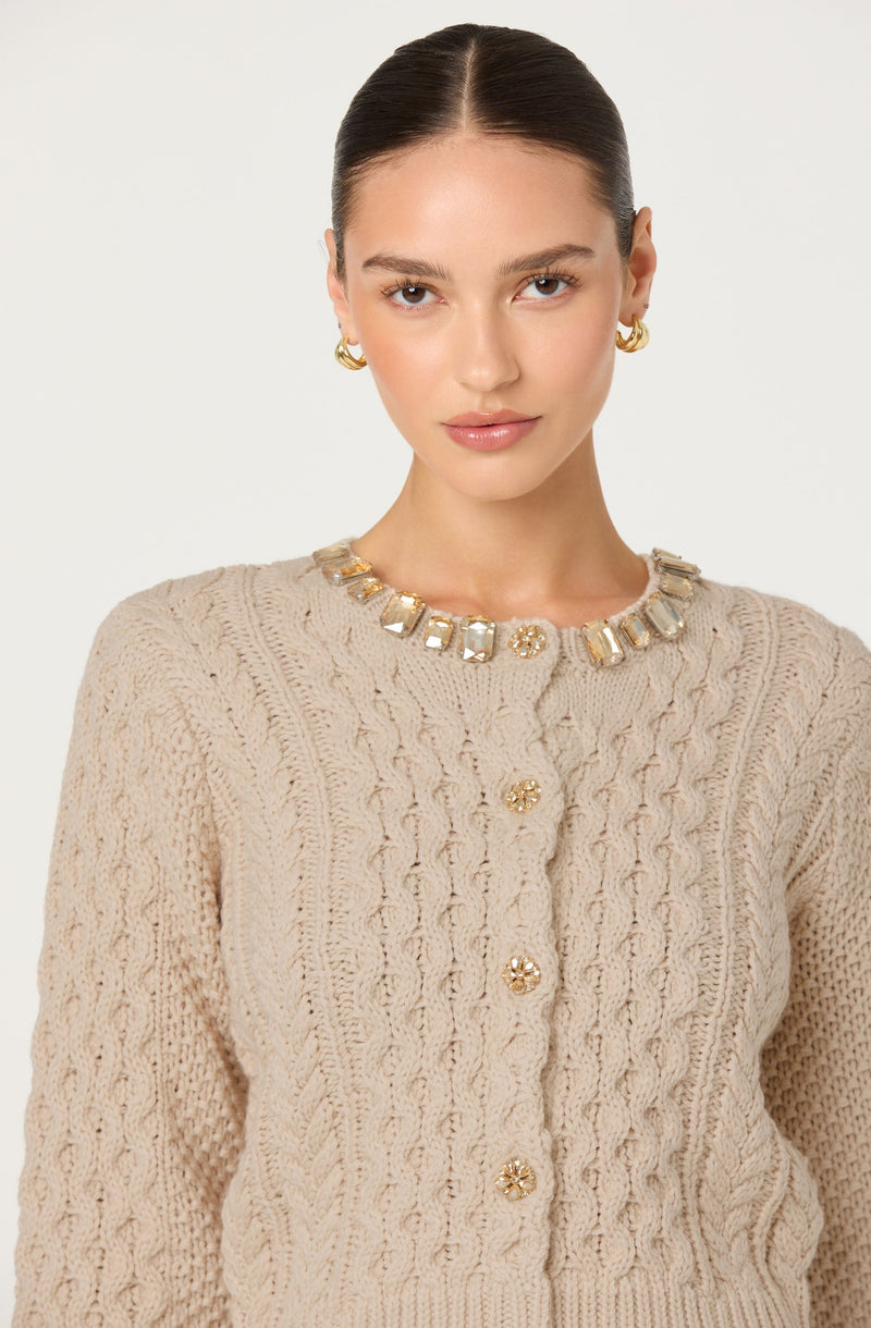 A woman is wearing a cream-colored, cable-knit sweater with a round neckline. The sweater features a textured pattern and has decorative gold buttons down the front. She is accessorized with a chunky necklace with large, square-shaped, clear stones and gold accents, and matching gold hoop earrings. She has her hair pulled back and has a neutral expression on her face.