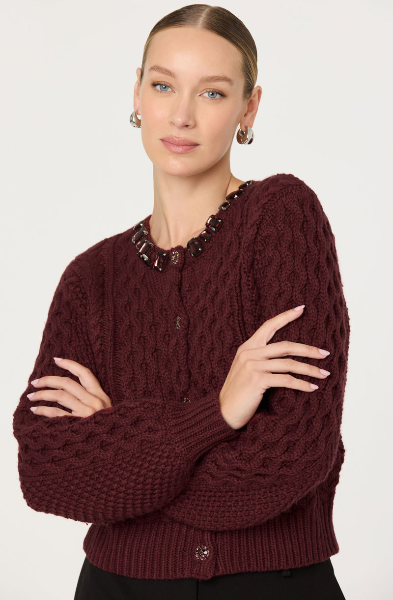 A woman is wearing a dark red, chunky knit sweater with cable patterns. The sweater has a rounded neckline decorated with multicolored beads and a small, subtle button closure at the front. The cuffs and hem of the sweater are ribbed.