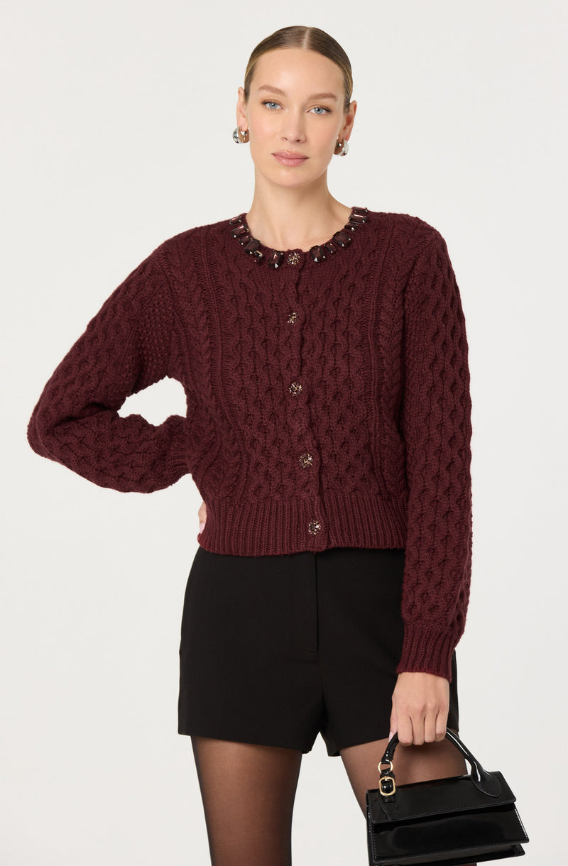 A woman is wearing a burgundy cable-knit cardigan with decorative embellishments around the neckline and buttons down the front. She pairs it with a black skirt and sheer black tights, and is holding a small black handbag in her left hand. She has her right hand on her hip and is looking directly at the camera.