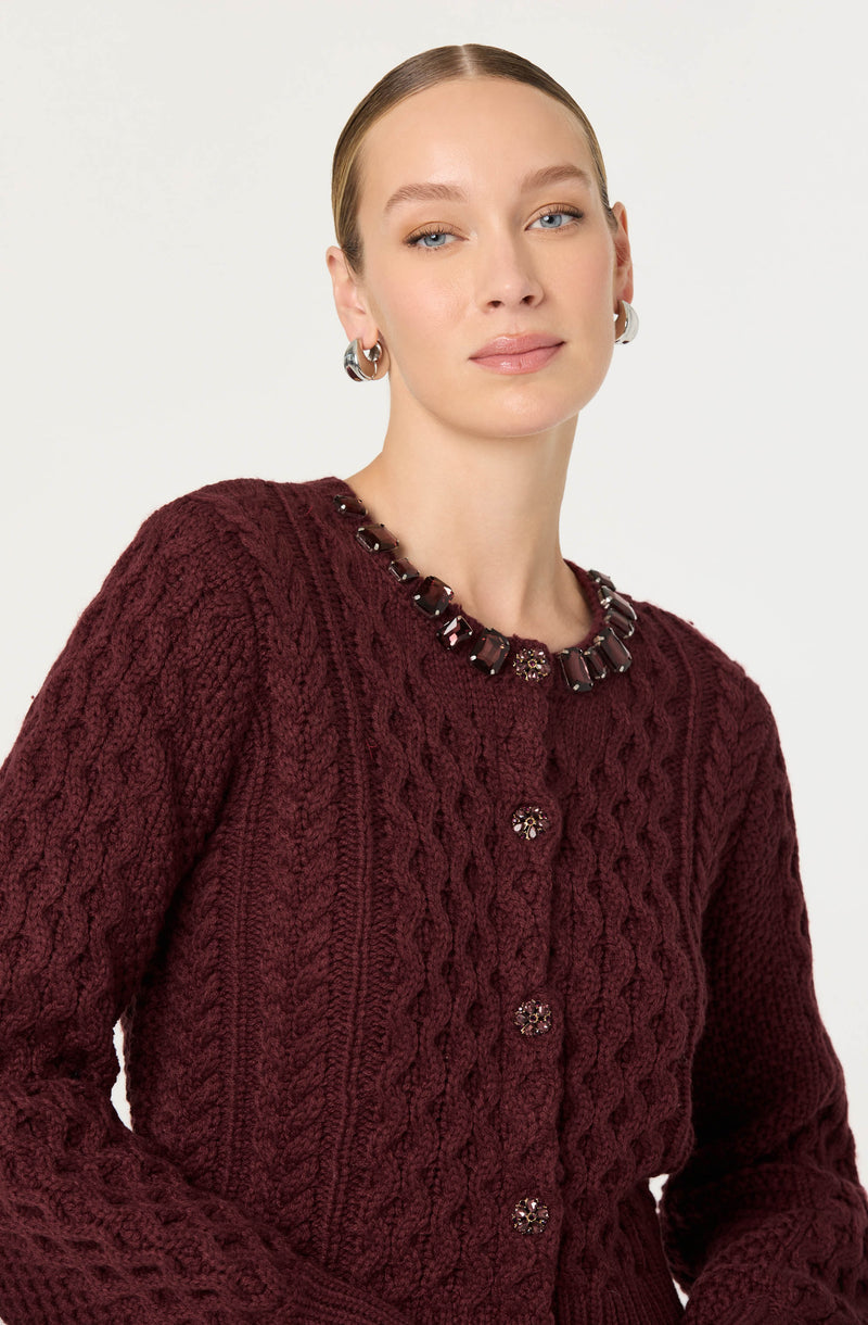 A woman wearing a burgundy cable-knit sweater with decorative buttons down the front. She is also accessorized with a dark beaded necklace and large silver hoop earrings.