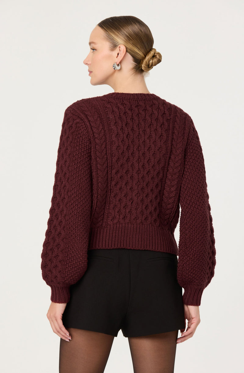 A woman is wearing a burgundy, chunky-knit sweater with cable and textured stitch patterns and balloon sleeves. She is also wearing black shorts and sheer black tights.