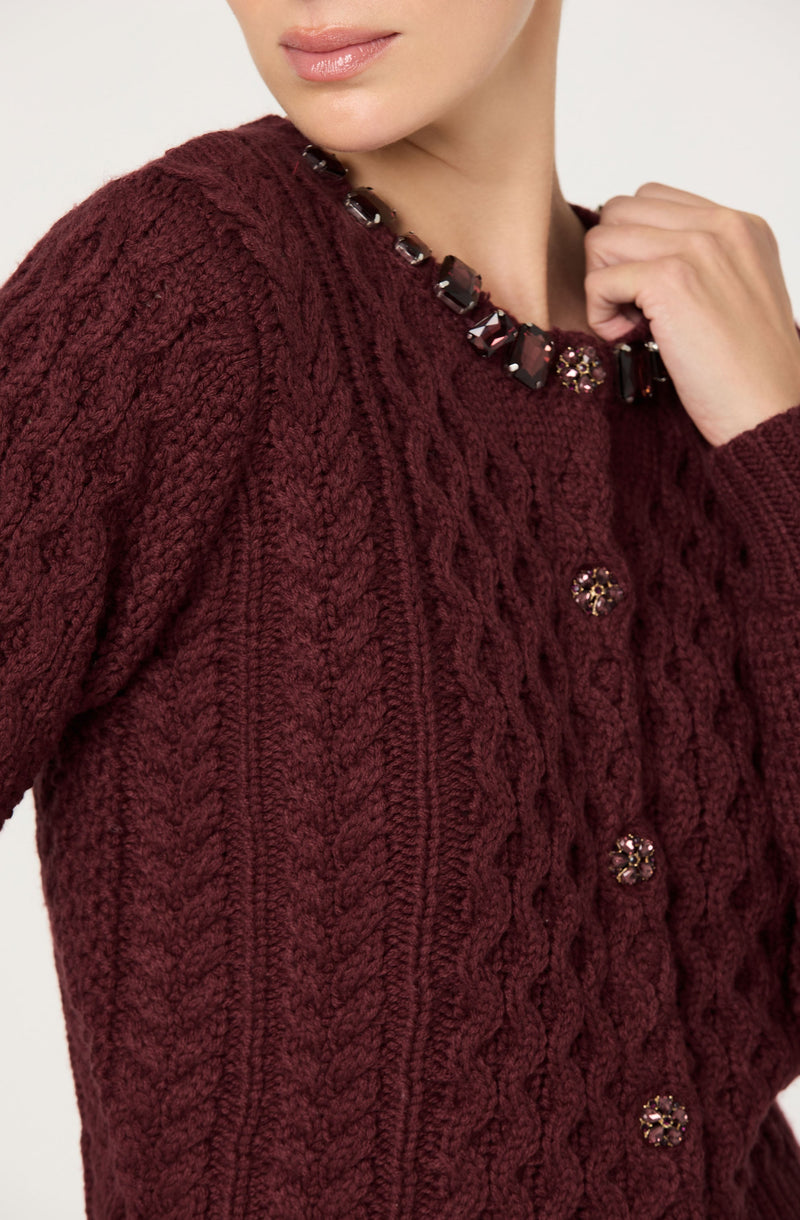 A woman is wearing a dark red, cable-knit sweater adorned with decorative buttons down the front, and a beaded necklace with large, dark, faceted stones and some smaller, sparkly embellishments.