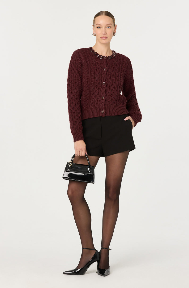 A woman is wearing a burgundy, textured knit cardigan with large buttons, paired with black high-waisted shorts. She has on sheer black tights and black high-heeled shoes with ankle straps. She is carrying a small, shiny black handbag with a gold chain handle.