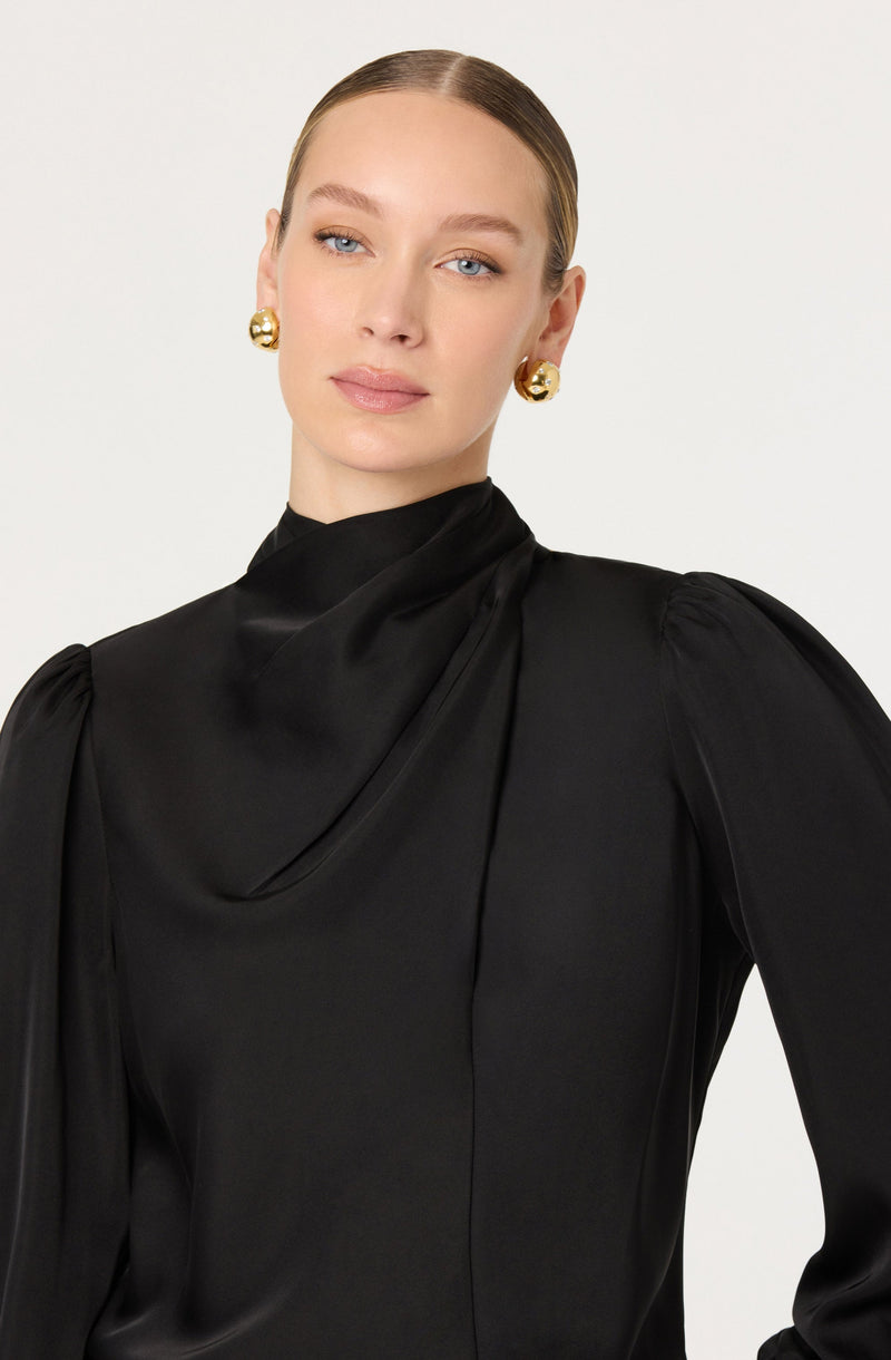 A woman is wearing a black satin top with a high neckline, featuring structured shoulders and puffed sleeves. She has accessorized with large, round gold earrings.