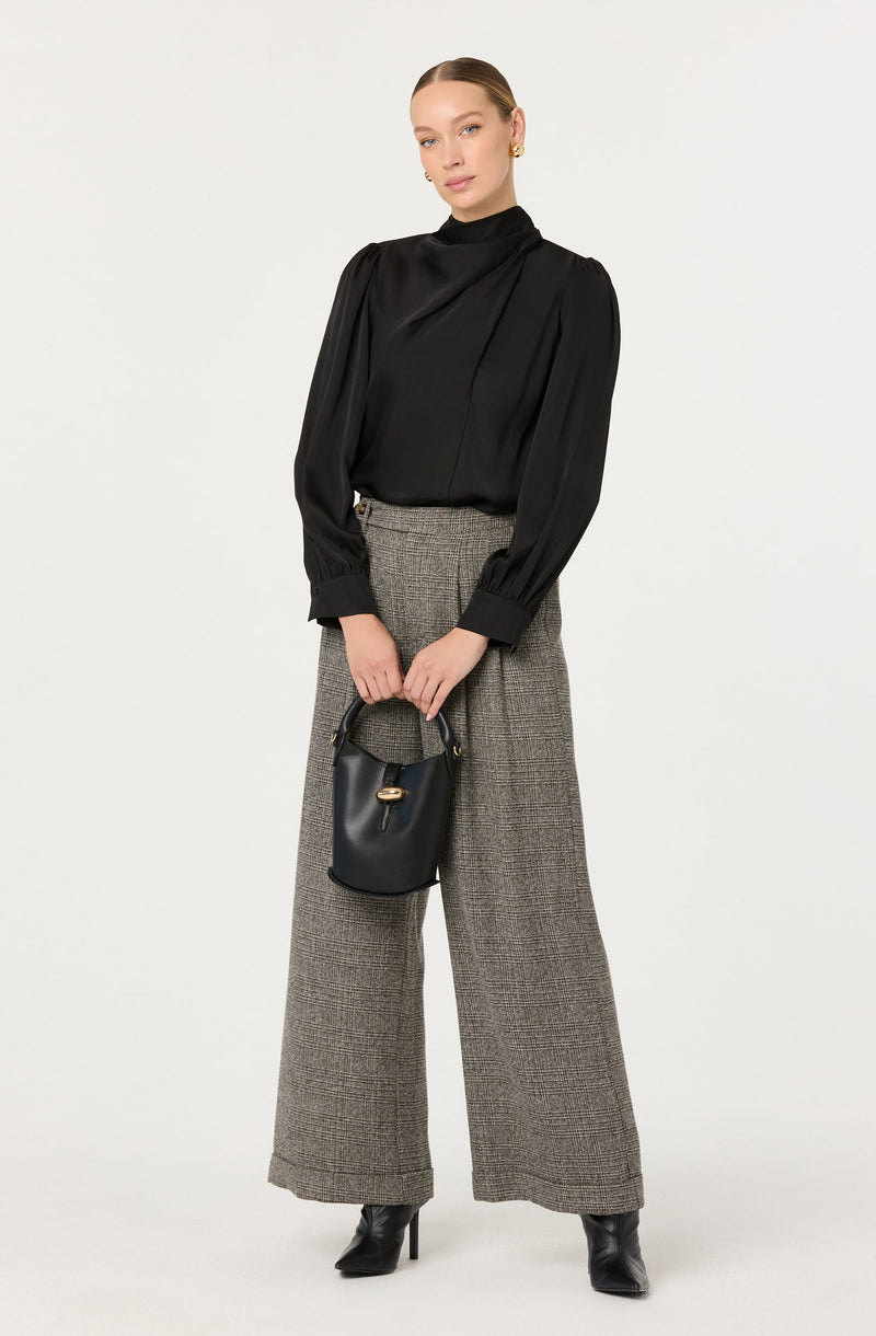 A woman is wearing a black long-sleeve blouse with a high neckline, featuring puffed sleeves gathered at the wrists. She pairs it with high-waisted, wide-leg trousers in a gray checkered pattern. She accessorizes with small gold earrings, a black handbag, and black high-heeled ankle boots.