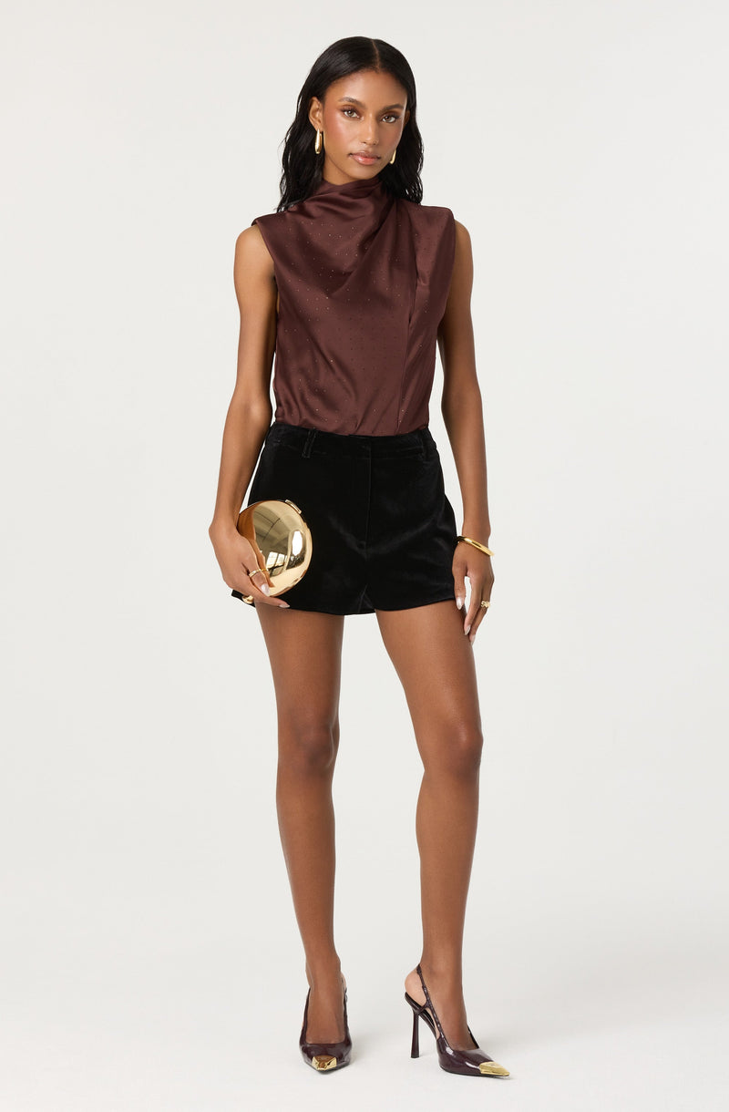 A woman is wearing a sleeveless, high-neck, silky brown top tucked into high-waisted black shorts. She accessorizes with gold hoop earrings, a gold bracelet, and a gold ring. She is holding a shiny gold clutch bag in her right hand and wears black high-heeled shoes with gold toe caps.