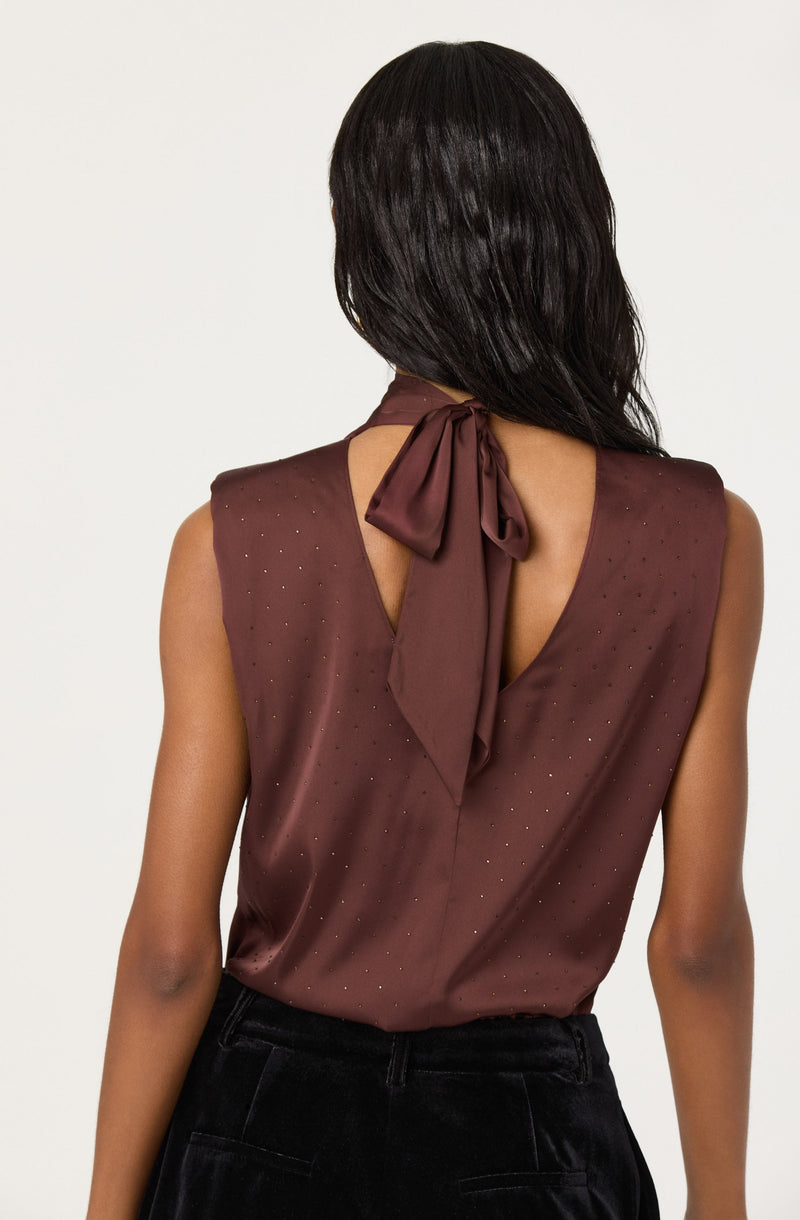 A woman is wearing a sleeveless, satin-like top in a rich brown color. The top features a subtle pattern of small, shiny dots scattered across the fabric. It has a deep V-shaped open back, with a matching brown satin bow tied at the neck, creating a stylish and elegant look.