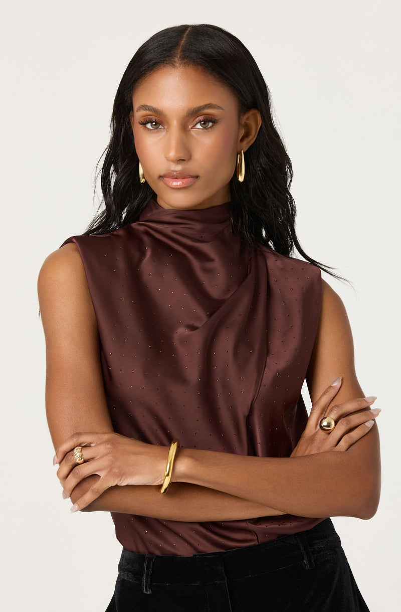 A woman is wearing a sleeveless, high-neck satin top in a deep burgundy color. The top has a subtle dotted pattern and a draped, slightly loose fit. She accessorizes with gold hoop earrings, a gold bracelet on her right wrist, a gold ring on her right middle finger, and a gold ring on her left index finger.