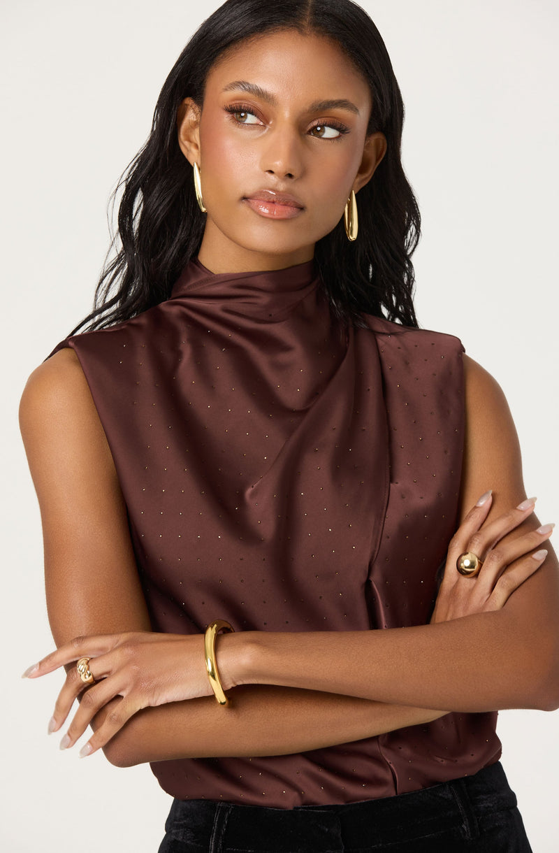 A woman is wearing a sleeveless, high-neck blouse made of shiny, satin-like fabric in a deep burgundy color. The blouse features tiny, subtle gold polka dots scattered across the fabric. She accessorizes with large gold hoop earrings, a gold bangle on her left wrist, a gold ring on her left index finger, and a gold statement ring on her right hand.