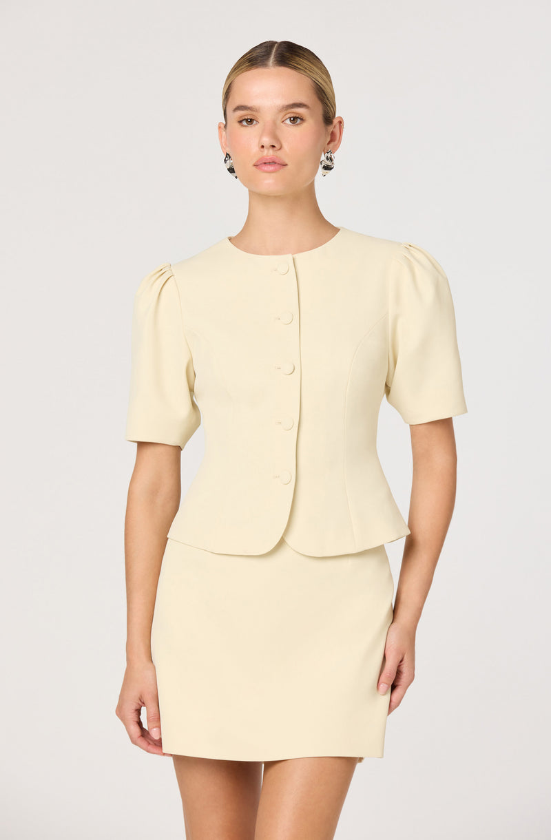 The model is wearing a cream-colored, short-sleeved top with puffed sleeves and a fitted, knee-length skirt that matches in color. The top has a rounded neckline, front button closure, and a slightly fitted waist. She is also wearing large, patterned earrings.