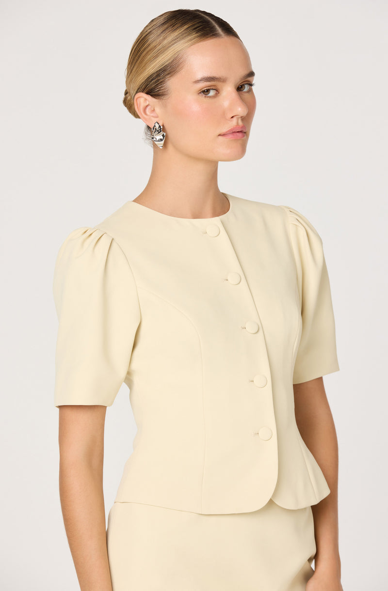 A woman is wearing a cream-colored, short-sleeved jacket with a rounded neckline and decorative pleats on the shoulders. The jacket features a row of fabric-covered buttons down the front. She complements her outfit with large, silver statement earrings.
