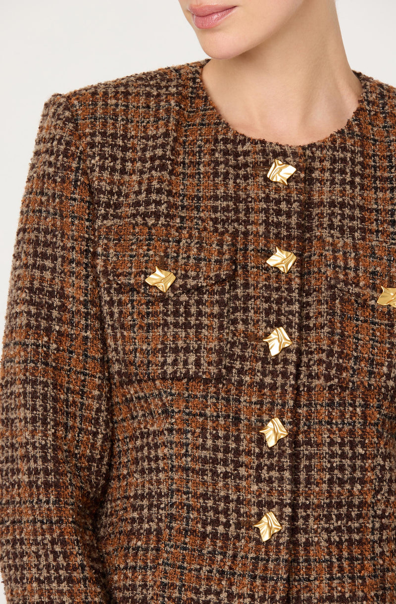 A woman is wearing a brown, plaid patterned jacket with a textured fabric. The jacket features a round neckline and is decorated with six metallic gold star-shaped buttons down the front. The plaid pattern includes shades of brown, black, and tan.