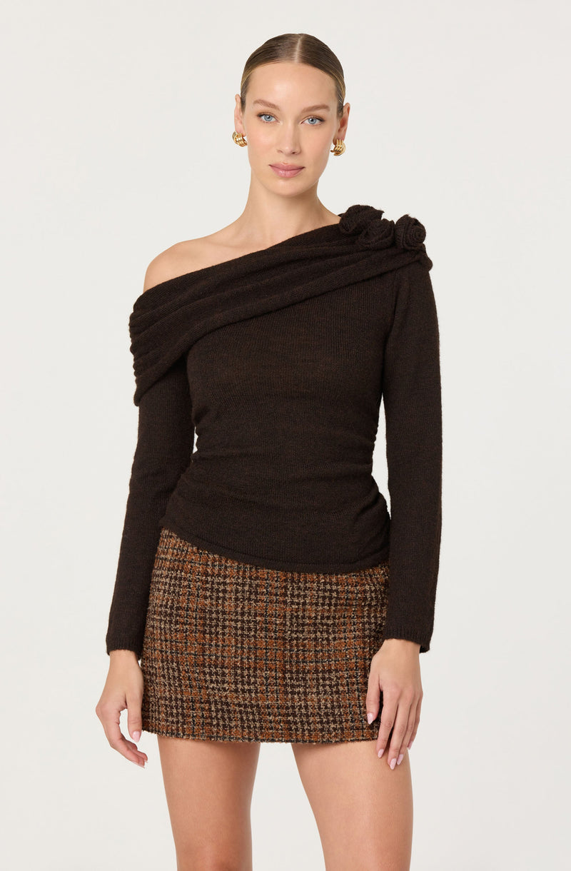 A woman is wearing a black off-the-shoulder sweater with a textured, twisted detail draped across her left shoulder. She pairs it with a short, fitted skirt featuring a brown and black checkered pattern. The overall look is stylish and sophisticated.
