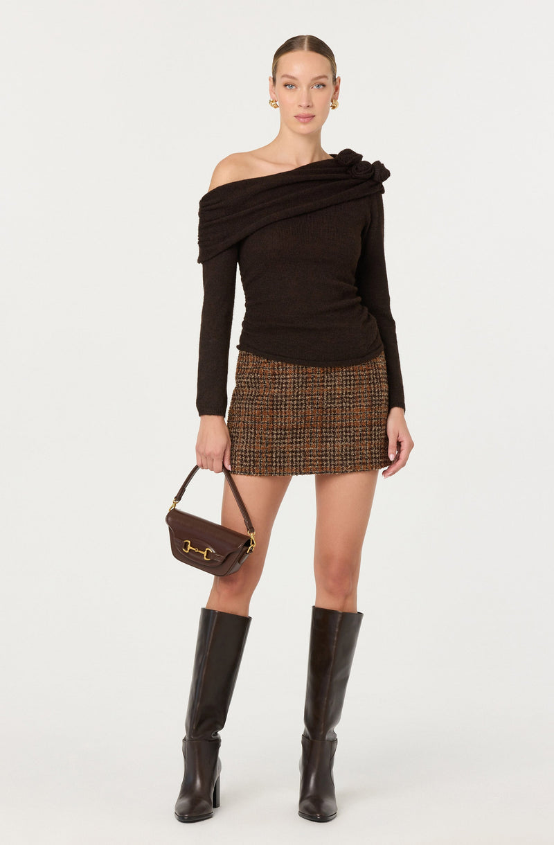 A woman is wearing a dark brown off-the-shoulder sweater with a large knot detail on the neckline. She pairs it with a high-waisted, short skirt in a brown, black, and orange checked pattern. She accessorizes with gold hoop earrings and carries a small brown handbag with gold hardware. She completes the outfit with tall, dark knee-high leather boots.