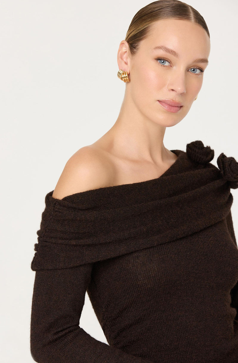 A woman with fair skin and blue eyes is wearing a dark brown off-the-shoulder sweater with a wide, ribbed, and slightly rolled neckline that forms a bow-like detail on her left side. She has gold, chunky, twisted hoop earrings and her hair is styled sleek and pulled back.