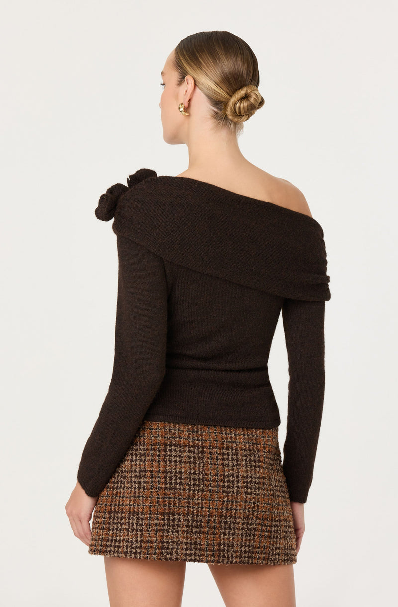 A woman is wearing a dark brown, off-the-shoulder sweater with a wide, draped collar that wraps around her shoulders and has a bow detail on the left side. She is also wearing a high-waisted, plaid mini skirt in shades of brown and beige.