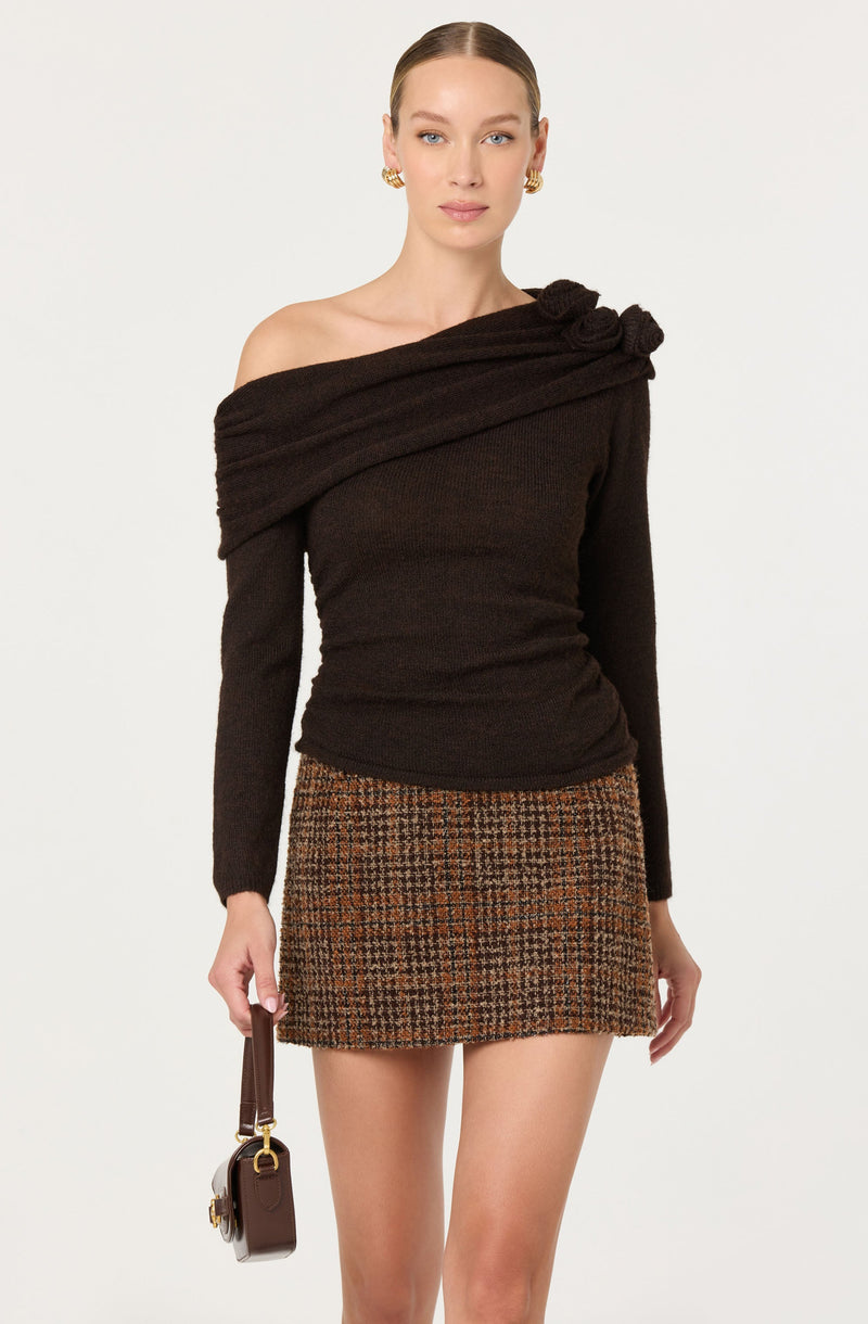 A woman is wearing a black, off-the-shoulder sweater with a textured, knotted detail on the shoulder. She pairs it with a brown, plaid mini skirt and gold hoop earrings. She is holding a small, dark brown handbag with a top handle.