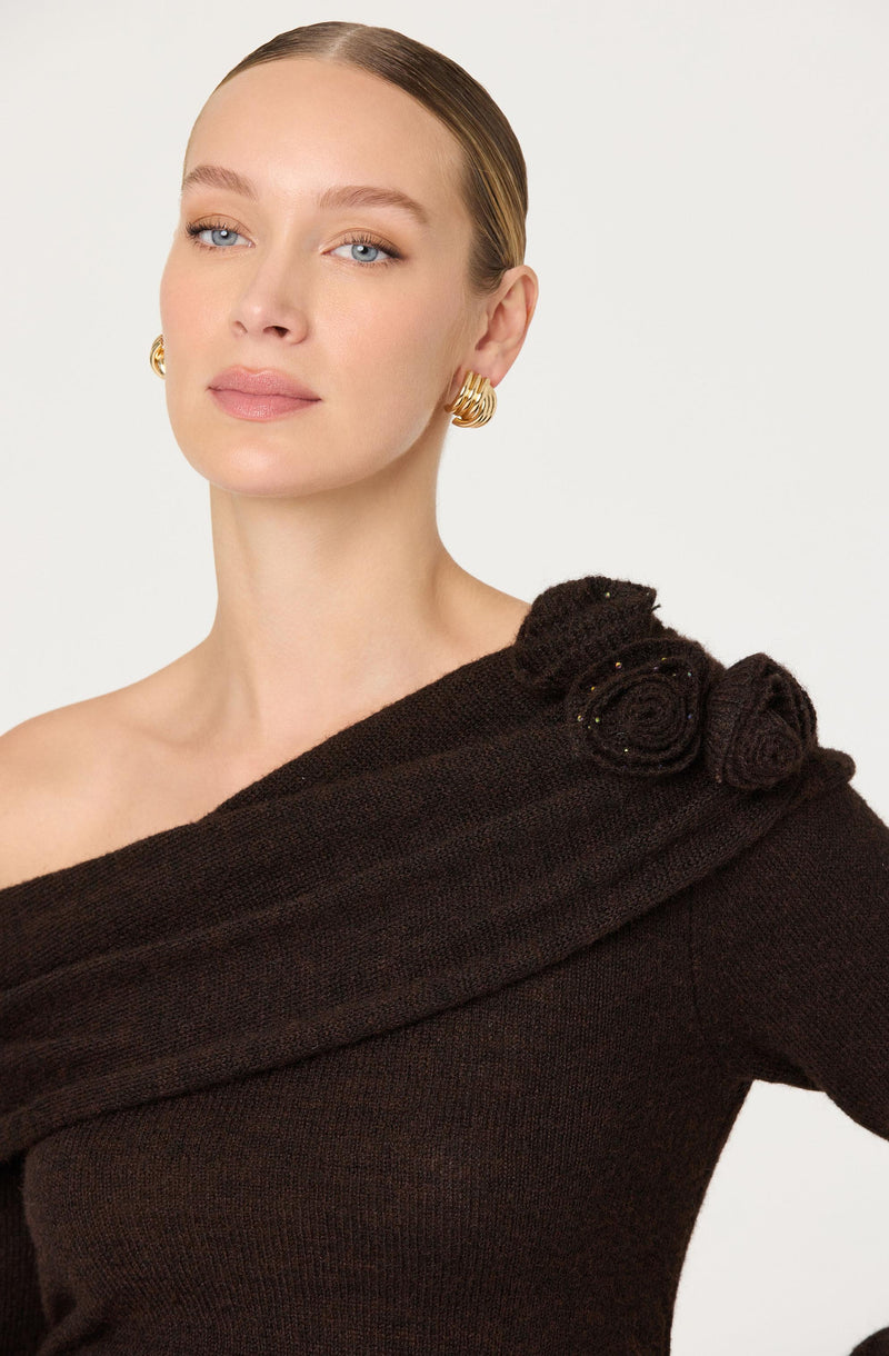 A woman with fair skin and light blue eyes is wearing a dark brown knit sweater with an off-the-shoulder design. The sweater features decorative knit rosettes on one shoulder. She has her hair styled in a sleek, pulled-back fashion and is accessorized with gold hoop earrings.