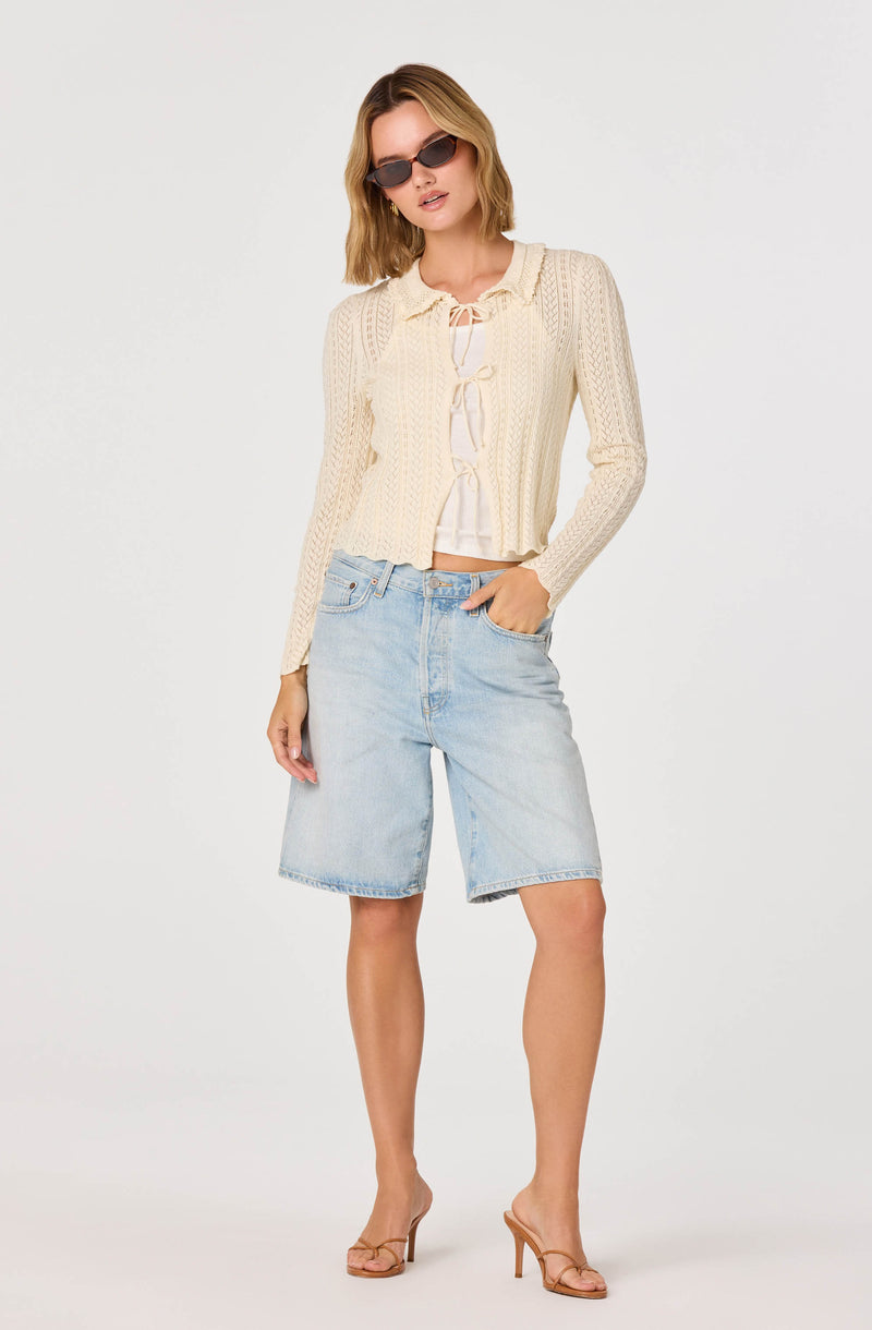 Woman in beige knit cardigan, white top, light denim shorts, and nude heels.