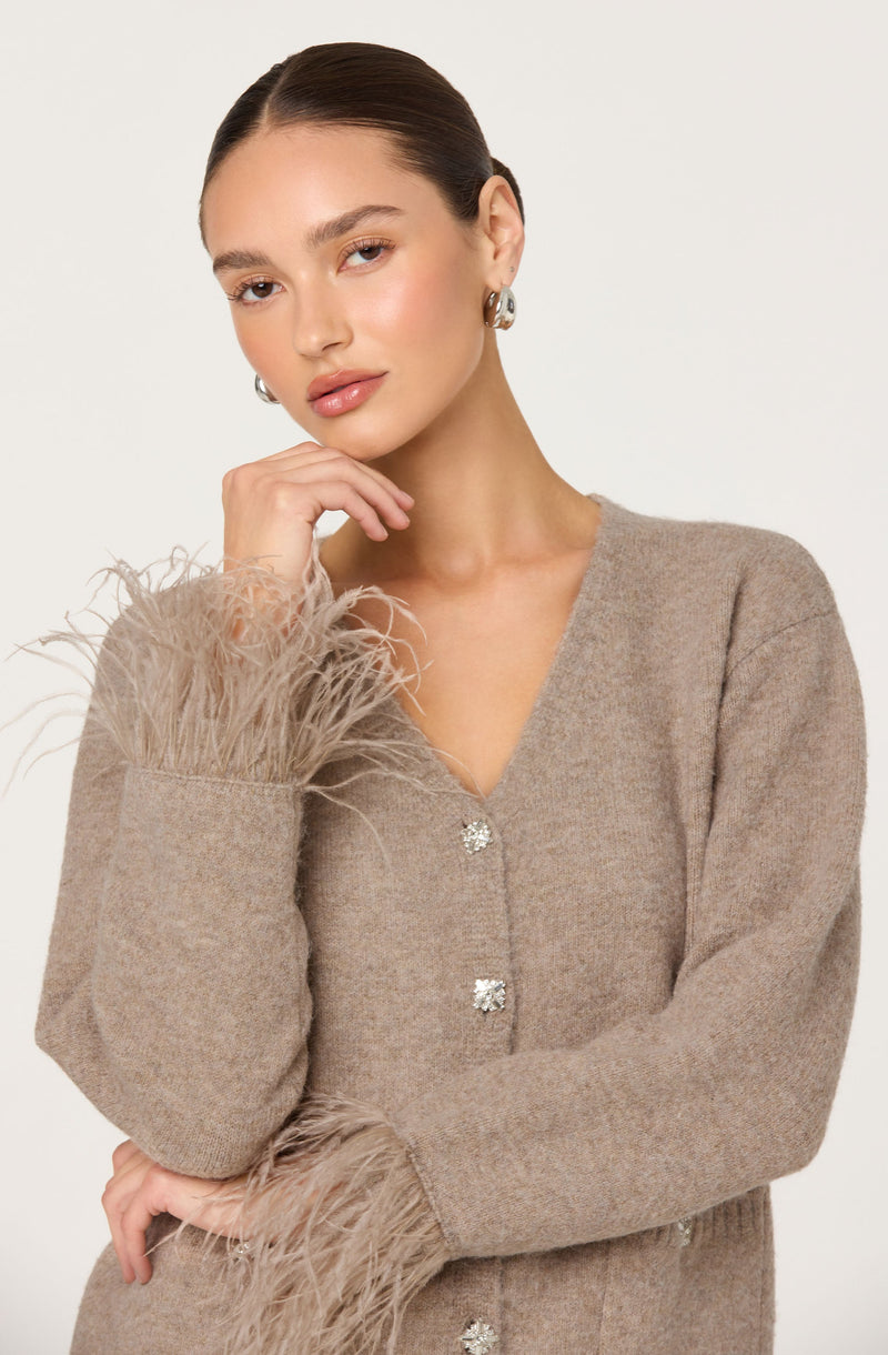 A woman is wearing a light brown, textured sweater with a V-neckline. The sweater features decorative buttons and has feather-like embellishments on the cuffs. She has her hair pulled back and is wearing large, shiny hoop earrings.