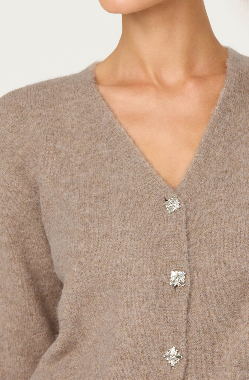 A woman is wearing a beige, fuzzy knit cardigan with a V-neckline. The cardigan has four decorative buttons that appear to be made of rhinestones or crystals arranged in flower shapes. The fabric looks soft and warm.