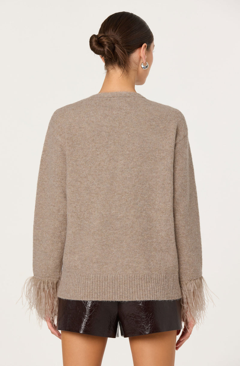 The woman is wearing a beige knit sweater with feathered cuffs at the sleeves, paired with shiny black shorts. She has her hair styled in a low bun and is accessorized with silver hoop earrings.