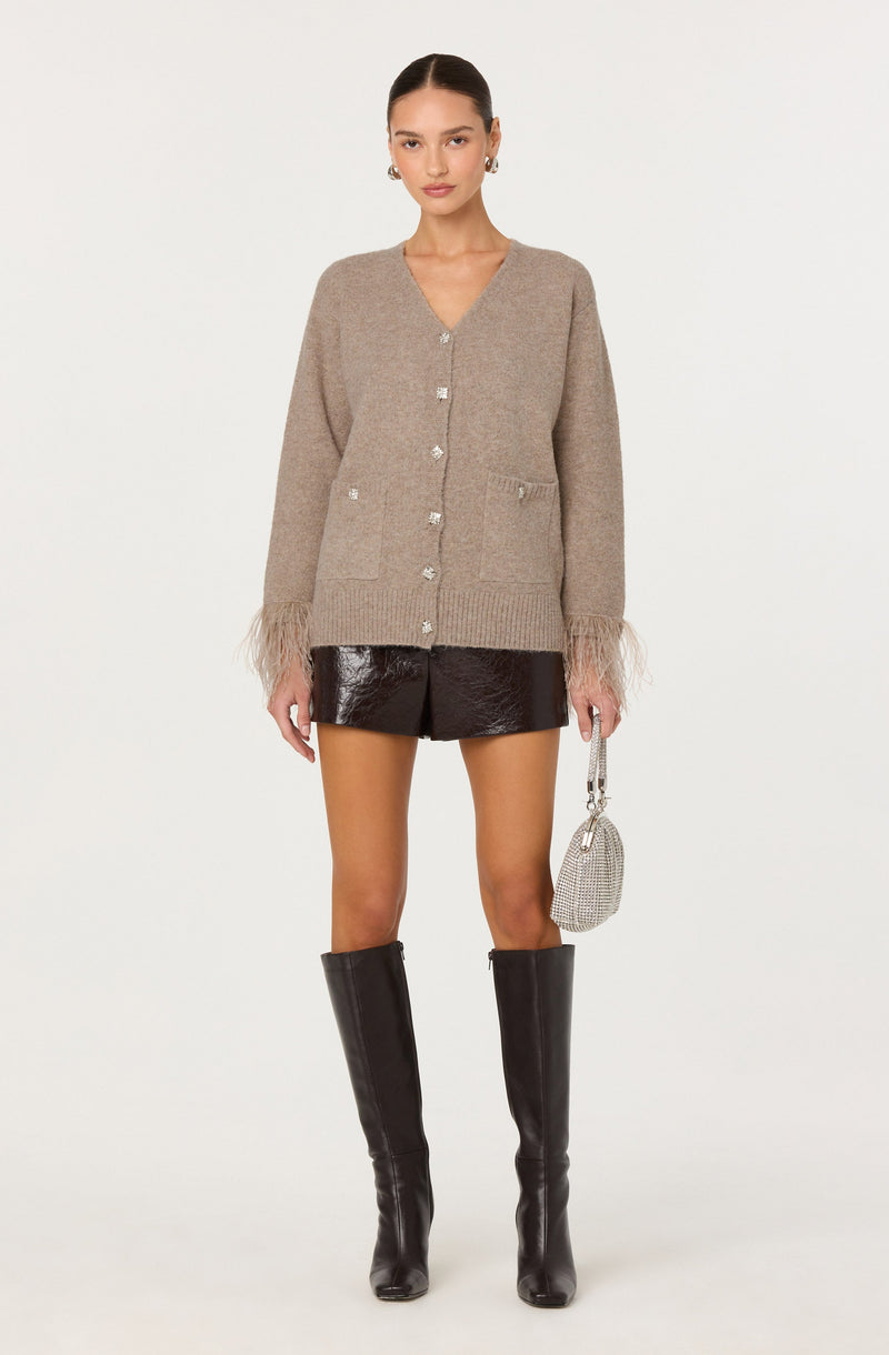 A woman is wearing a beige, oversized cardigan with a V-neckline, button closure down the front, and two front pockets. The cardigan features fringe details at the cuffs. She is also dressed in shiny black shorts and tall black knee-high boots. She is holding a small, silver, rhinestone-embellished handbag.