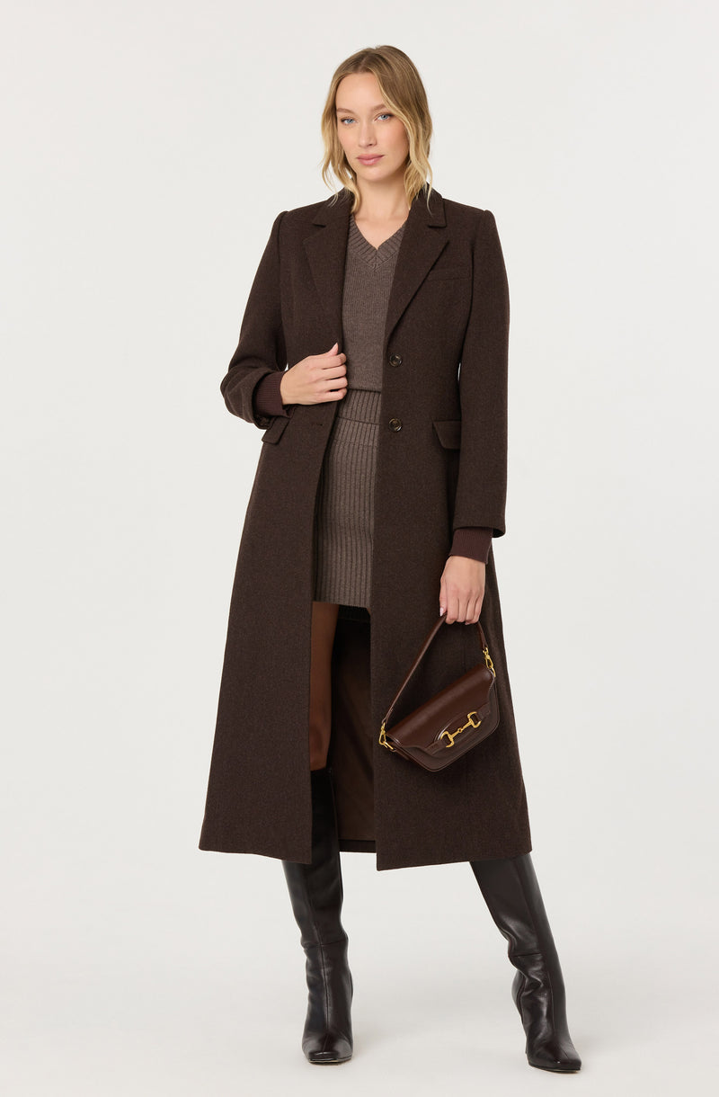 Woman in a long dark brown coat, brown sweater, and knee-high black boots, holding a brown handbag.