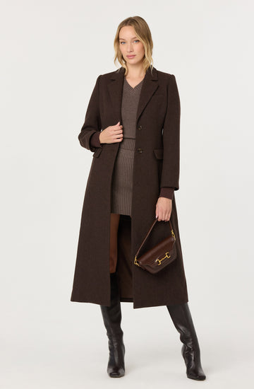 Woman in a long dark brown coat, brown sweater, and knee-high black boots, holding a brown handbag.
