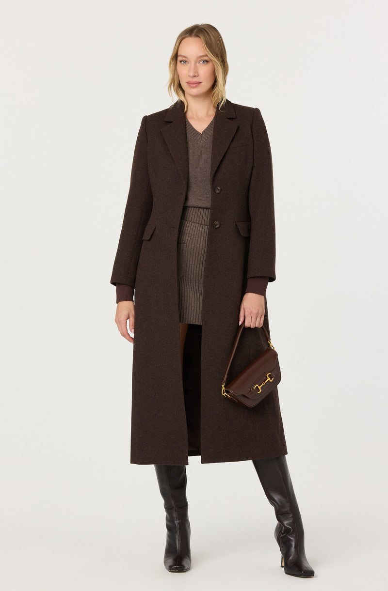 Woman in a long dark brown coat, gray sweater, matching skirt, and black knee-high boots, holding a brown handbag.