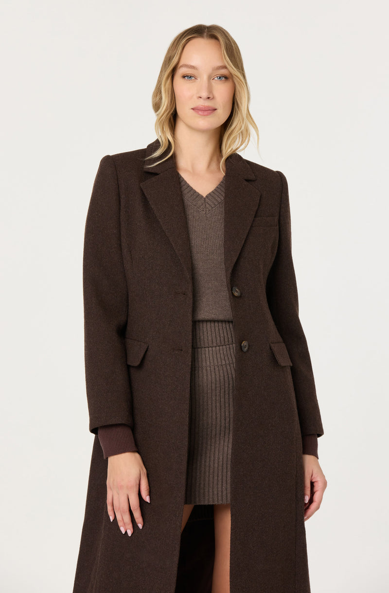 Woman wearing a long brown coat over a matching brown sweater and ribbed skirt.