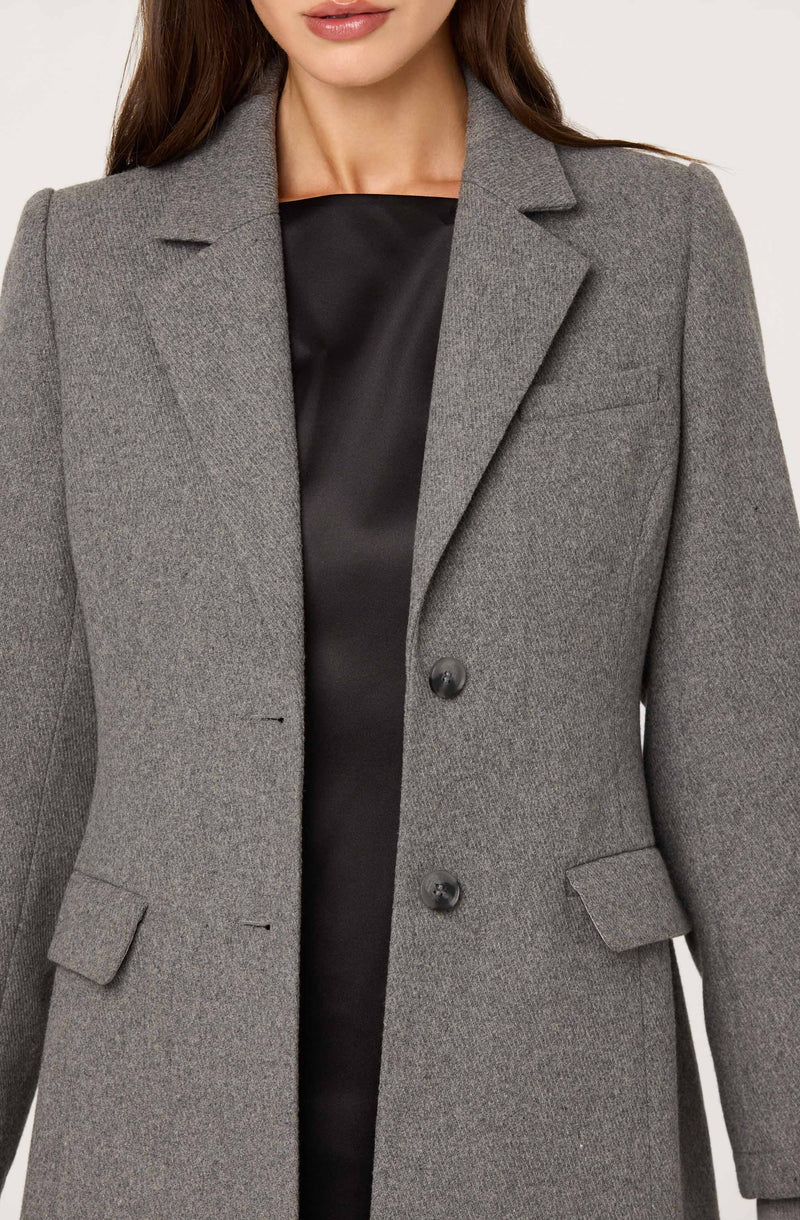 A woman is wearing a tailored gray blazer with notch lapels, two large buttons down the front, and flap pockets on each side. She is also wearing a black satin top underneath the blazer.