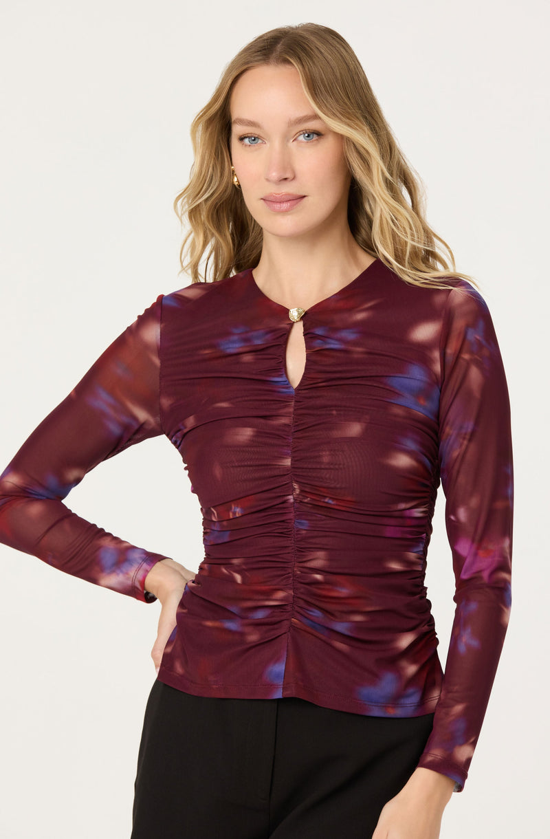 A woman is wearing a long-sleeved, fitted top with a maroon base color accented by blue and purple tie-dye patterns. The top features ruching down the center front, ending at a small keyhole opening closed with a decorative button.
