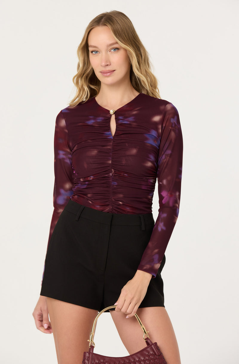 A woman is wearing a long-sleeved, fitted top with a keyhole neckline and a small decorative button. The top features a dark, burgundy base color with a purple and blue tie-dye pattern. She pairs it with high-waisted black shorts and holds a small, woven burgundy handbag.