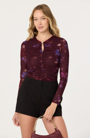 A woman is wearing a long-sleeved, fitted top with a keyhole neckline and a small decorative button. The top features a dark, burgundy base color with a purple and blue tie-dye pattern. She pairs it with high-waisted black shorts and holds a small, woven burgundy handbag.