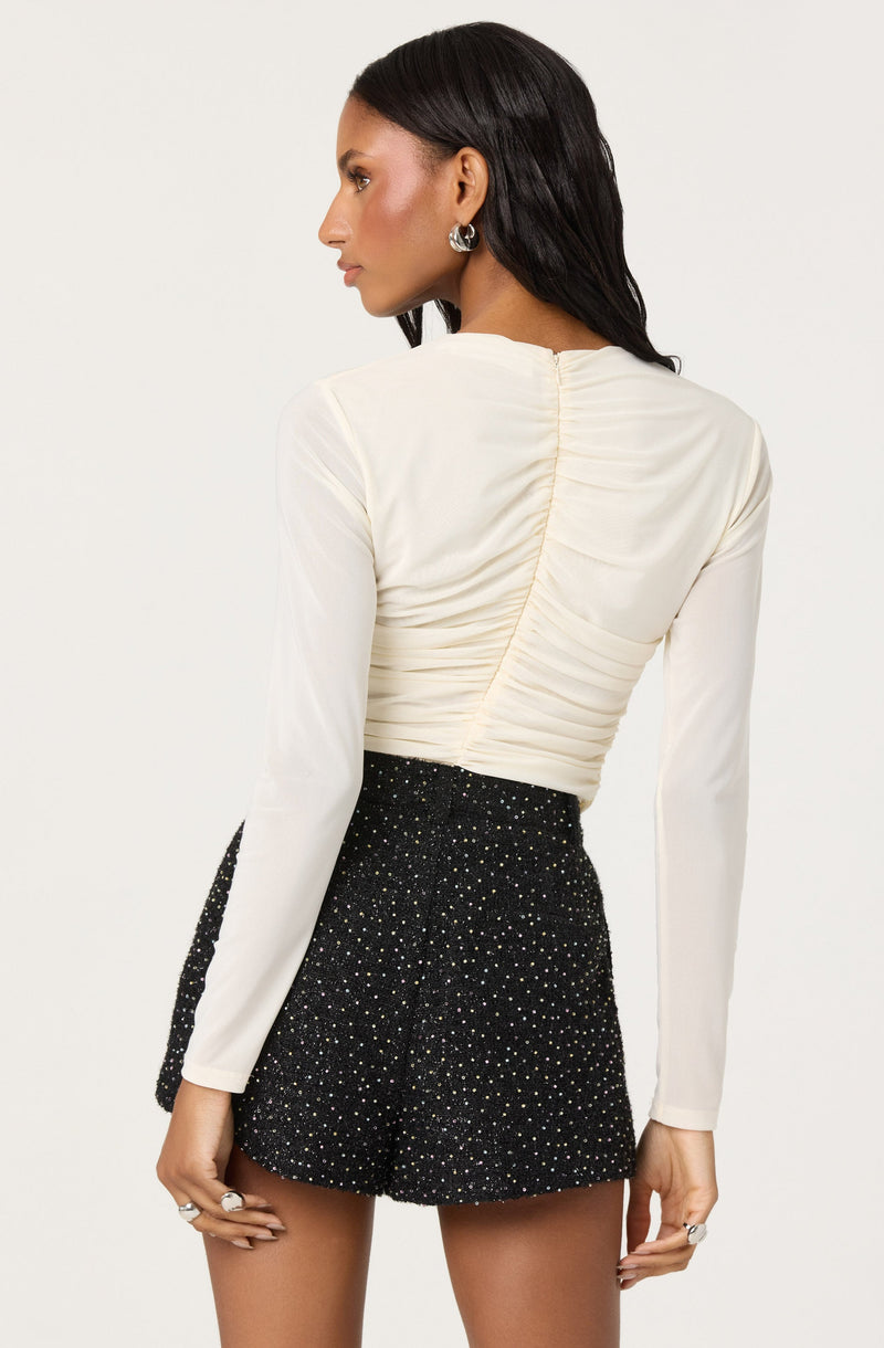 A woman is wearing a long-sleeved, cream-colored top with ruching details along the back. She pairs it with high-waisted black shorts that are decorated with multicolored specks or rhinestones, creating a sparkling effect.
